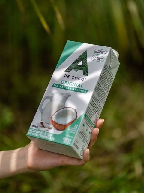Una mano sostiene Néctar Original A de Coco frente a hojas verdes, de @adecocomx